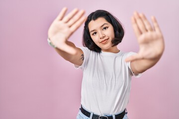 Young hispanic woman wearing casual white t shirt over pink background doing frame using hands palms and fingers, camera perspective