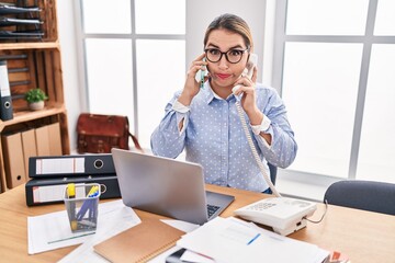 Young hispanic business woman working at the office talking on two phones puffing cheeks with funny face. mouth inflated with air, catching air. - obrazy, fototapety, plakaty