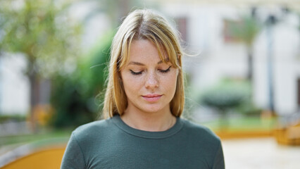 Young blonde woman standing with serious expression looking down at park