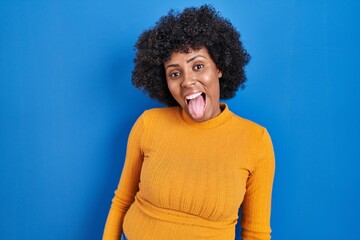 Fototapeta na wymiar Black woman with curly hair standing over blue background sticking tongue out happy with funny expression. emotion concept.