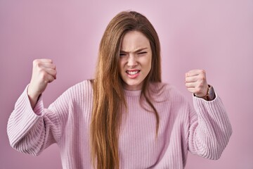 Young caucasian woman standing over pink background angry and mad raising fists frustrated and...