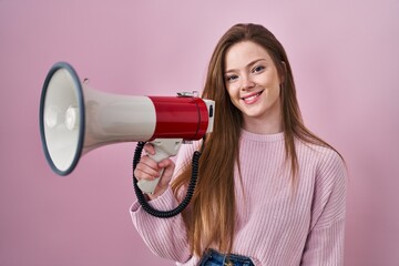 Young caucasian woman shouting through megaphone looking positive and happy standing and smiling with a confident smile showing teeth