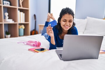 Young latin woman using laptop and credit card lying on bed at bedroom