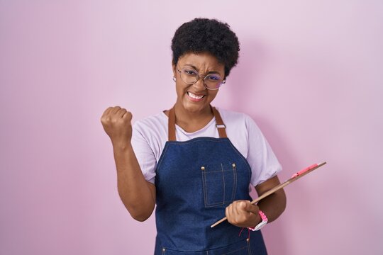 Young african american woman wearing professional waitress apron holding clipboard very happy and excited doing winner gesture with arms raised, smiling and screaming for success. celebration concept.