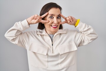 Middle age hispanic woman standing over isolated background doing peace symbol with fingers over face, smiling cheerful showing victory