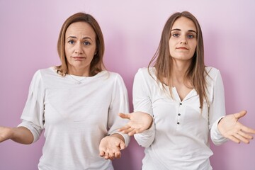 Middle age mother and young daughter standing over pink background clueless and confused with open arms, no idea concept.