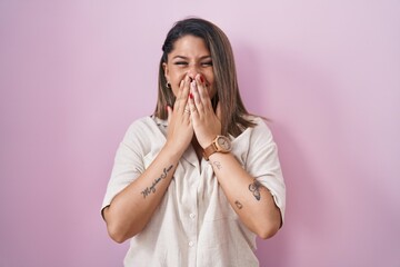 Blonde woman standing over pink background laughing and embarrassed giggle covering mouth with hands, gossip and scandal concept