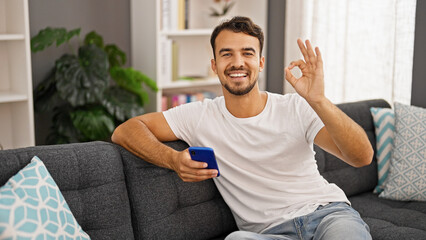 Young hispanic man using smartphone doing ok gesture at home
