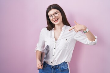 Young caucasian woman holding laptop looking confident with smile on face, pointing oneself with fingers proud and happy.