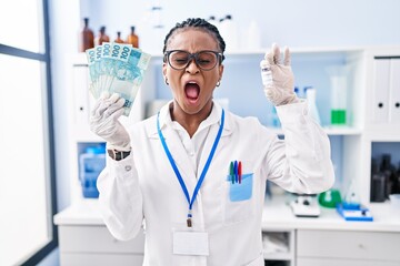 African woman with braids working at scientist laboratory holding money angry and mad screaming frustrated and furious, shouting with anger. rage and aggressive concept.