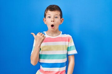 Young caucasian kid standing over blue background surprised pointing with hand finger to the side, open mouth amazed expression.