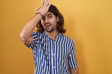 Hispanic man with long hair standing over yellow background surprised with hand on head for mistake, remember error. forgot, bad memory concept.
