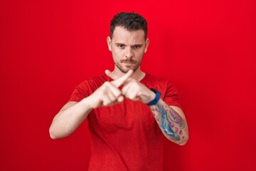 Young hispanic man standing over red background rejection expression crossing fingers doing negative sign - obrazy, fototapety, plakaty