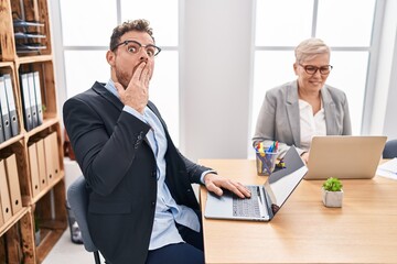 Hispanic young man working at the office covering mouth with hand, shocked and afraid for mistake....