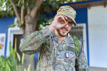 Young hispanic man wearing camouflage army uniform outdoors smiling happy doing ok sign with hand on eye looking through fingers