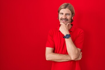 Caucasian man with mustache standing over red background looking confident at the camera with smile with crossed arms and hand raised on chin. thinking positive.