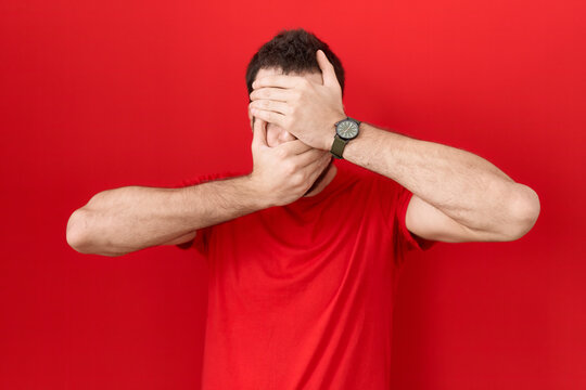 Young hispanic man wearing casual red t shirt covering eyes and mouth with hands, surprised and shocked. hiding emotion
