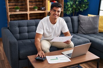 Young hispanic man using laptop reading document accounting at home