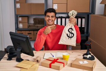 Hispanic man working at small business ecommerce holding money bag smiling happy pointing with hand and finger