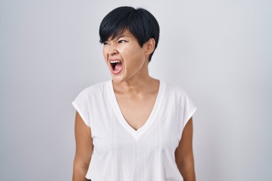 Young asian woman with short hair standing over isolated background angry and mad screaming frustrated and furious, shouting with anger. rage and aggressive concept.