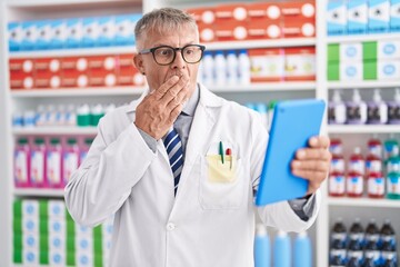 Hispanic man with grey hair working at pharmacy drugstore doing video call with tablet covering mouth with hand, shocked and afraid for mistake. surprised expression