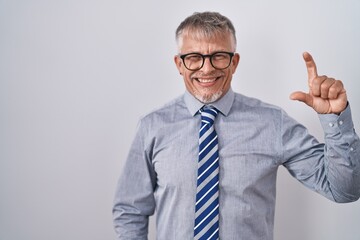 Hispanic business man with grey hair wearing glasses smiling and confident gesturing with hand doing small size sign with fingers looking and the camera. measure concept.
