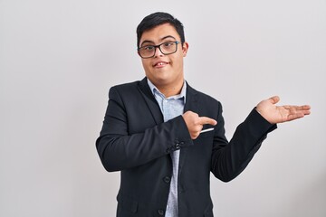 Obraz na płótnie Canvas Young hispanic man with down syndrome wearing business style amazed and smiling to the camera while presenting with hand and pointing with finger.