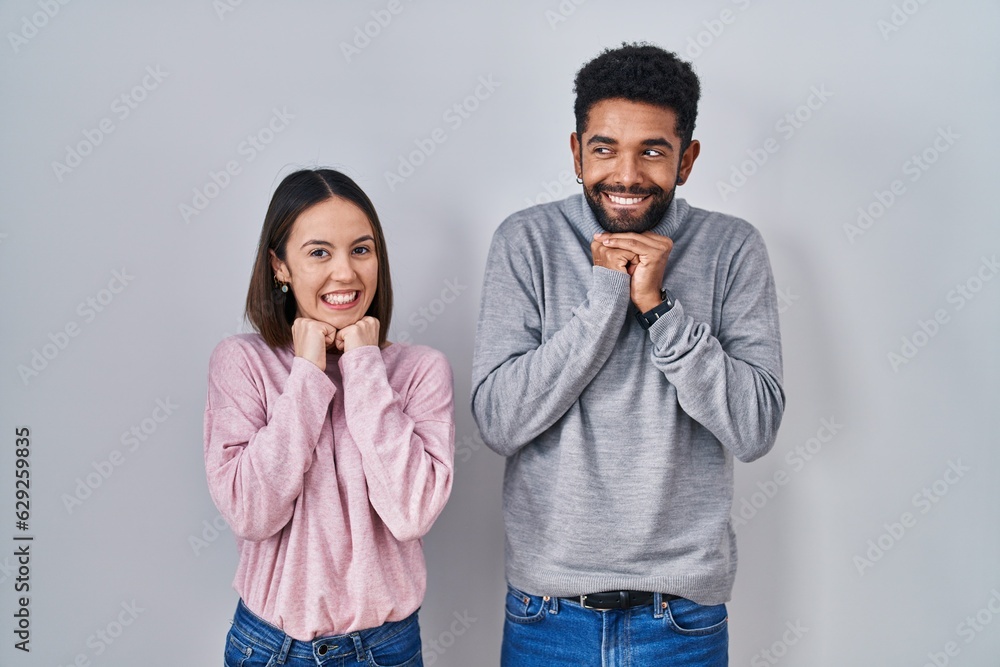 Wall mural Young hispanic couple standing together laughing nervous and excited with hands on chin looking to the side