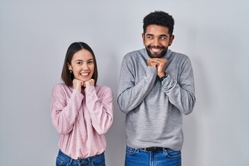 Young hispanic couple standing together laughing nervous and excited with hands on chin looking to the side