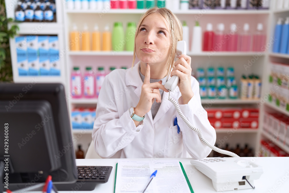 Canvas Prints Young caucasian woman working at pharmacy drugstore speaking on the telephone thinking concentrated about doubt with finger on chin and looking up wondering