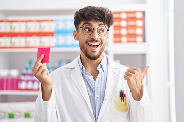 Arab man with beard working at pharmacy drugstore holding condom winking looking at the camera with...