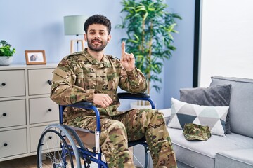 Arab man wearing camouflage army uniform sitting on wheelchair showing and pointing up with finger number one while smiling confident and happy.