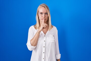 Young caucasian woman standing over blue background asking to be quiet with finger on lips. silence and secret concept.