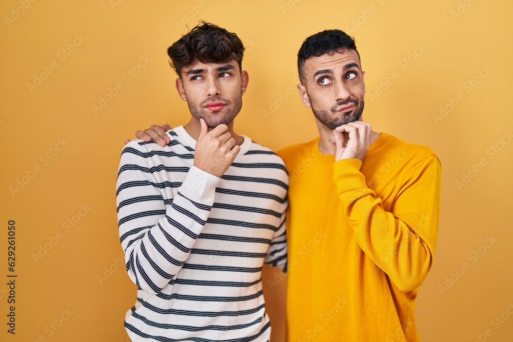 Wall mural Young hispanic gay couple standing over yellow background with hand on chin thinking about question, pensive expression. smiling with thoughtful face. doubt concept.