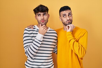 Young hispanic gay couple standing over yellow background with hand on chin thinking about question, pensive expression. smiling with thoughtful face. doubt concept.