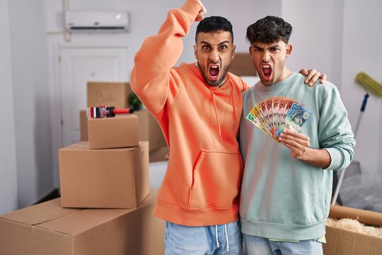 Young Hispanic Gay Couple Moving To A New Home Holding Banknotes Annoyed And Frustrated Shouting With Anger, Yelling Crazy With Anger And Hand Raised