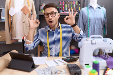 Hispanic man with beard dressmaker designer working at atelier looking surprised and shocked doing ok approval symbol with fingers. crazy expression