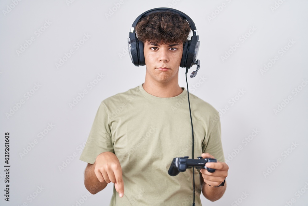 Wall mural hispanic teenager playing video game holding controller pointing down looking sad and upset, indicat