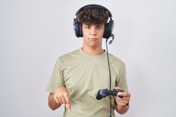 Hispanic teenager playing video game holding controller pointing down looking sad and upset,...