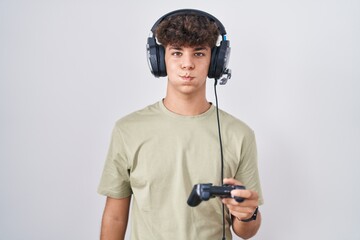 Hispanic teenager playing video game holding controller puffing cheeks with funny face. mouth...