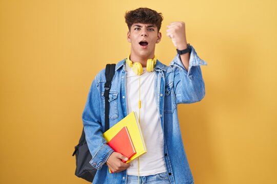 Hispanic teenager wearing student backpack and holding books angry and mad raising fist frustrated and furious while shouting with anger. rage and aggressive concept.