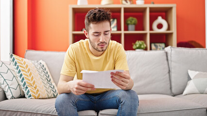 Young hispanic man reading document sitting on sofa with serious face at home