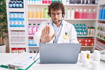 Hispanic young man working at pharmacy drugstore working with laptop with open hand doing stop sign with serious and confident expression, defense gesture