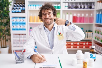 Hispanic young man working at pharmacy drugstore pointing with hand finger to face and nose, smiling cheerful. beauty concept