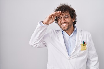 Hispanic young man wearing doctor uniform very happy and smiling looking far away with hand over head. searching concept.