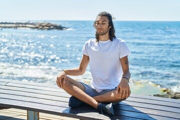 Young hispanic man doing yoga exercise sitting on bench at seaside