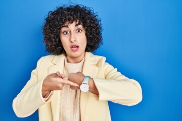 Young brunette woman with curly hair standing over blue background in hurry pointing to watch time,...