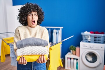 Young brunette woman with curly hair holding clean laundry angry and mad screaming frustrated and...