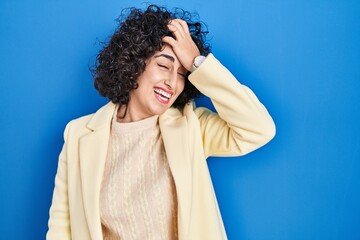 Young brunette woman with curly hair standing over blue background surprised with hand on head for mistake, remember error. forgot, bad memory concept.