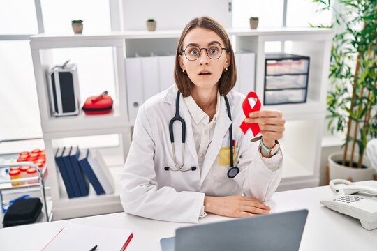 Young caucasian doctor woman holding support red ribbon scared and amazed with open mouth for surprise, disbelief face
